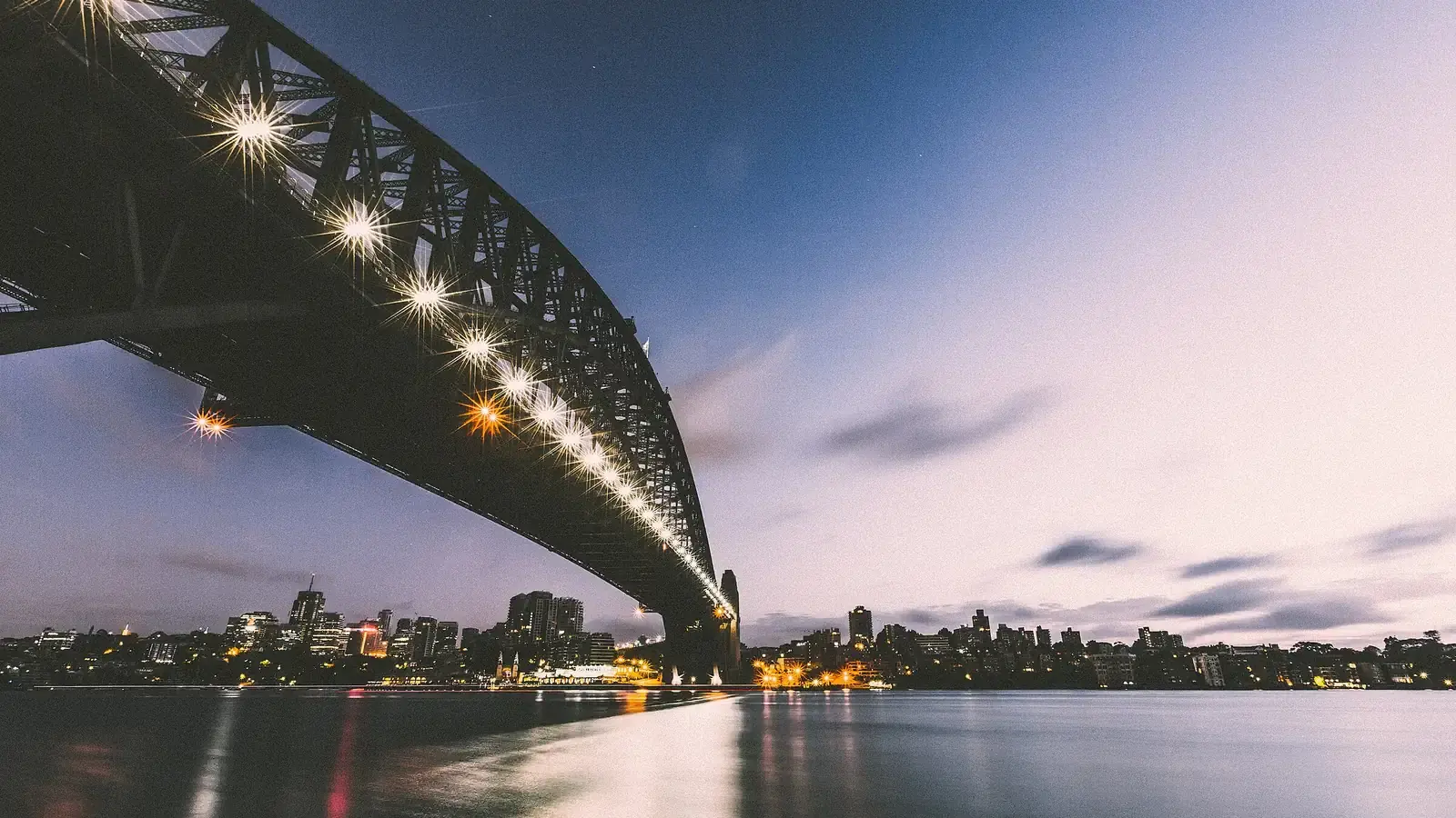 Sydney Harbour Bridge lit at dusk, the city skyline low on the far shore