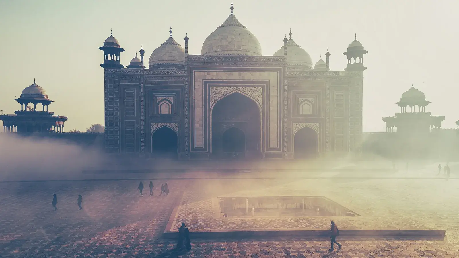 The marble domes of the Taj Mahal complex through morning mist, figures crossing the courtyard