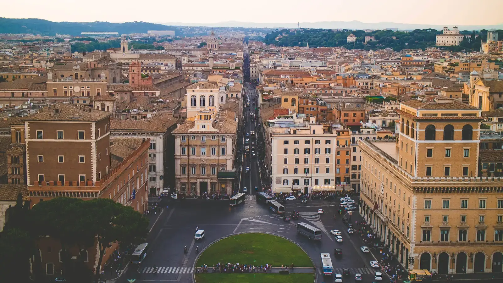 Rome's rooftops at dusk from above, a straight avenue running back from a busy square