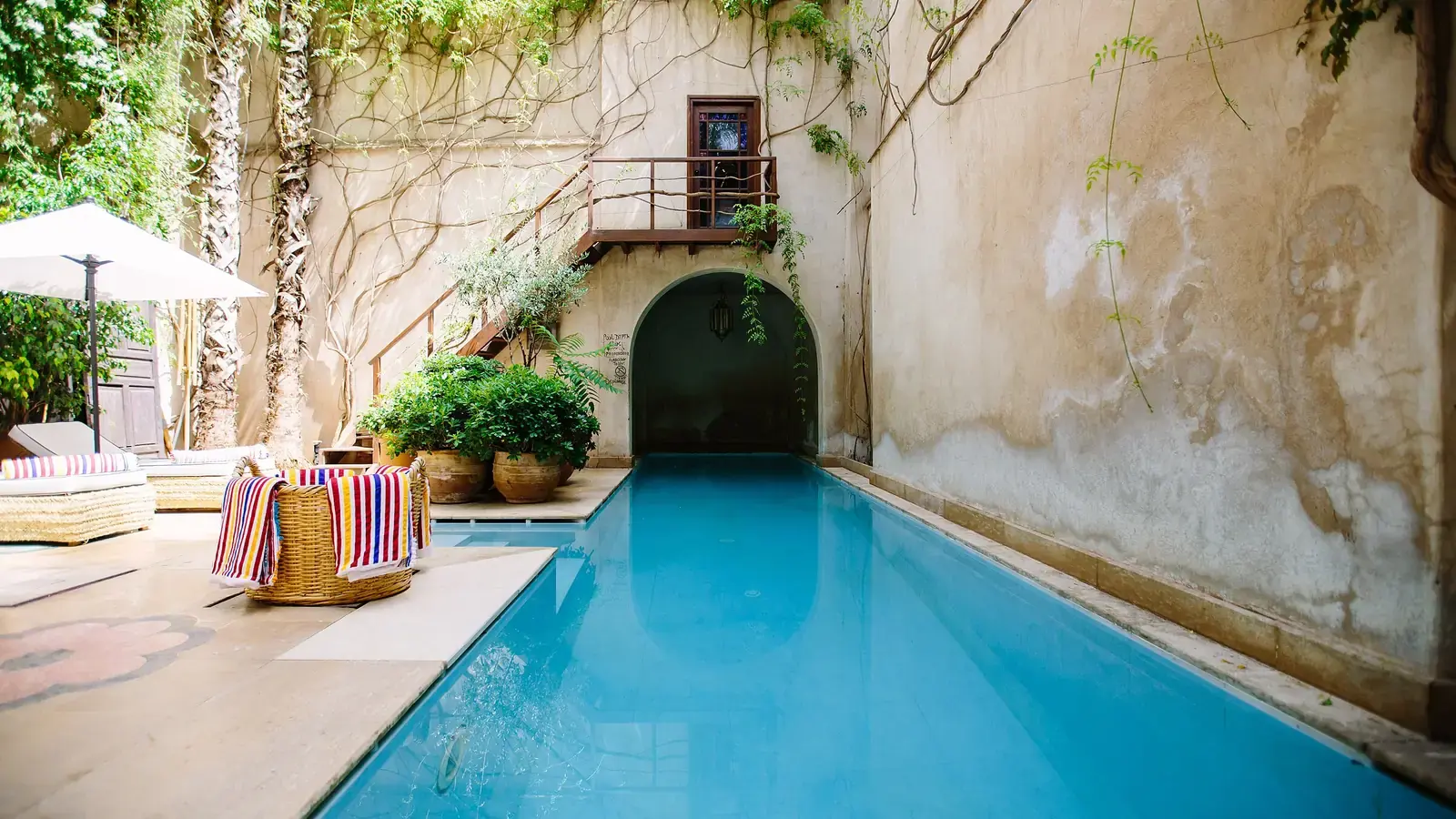 A long pool in a riad courtyard, ochre walls and striped towels over a wicker basket