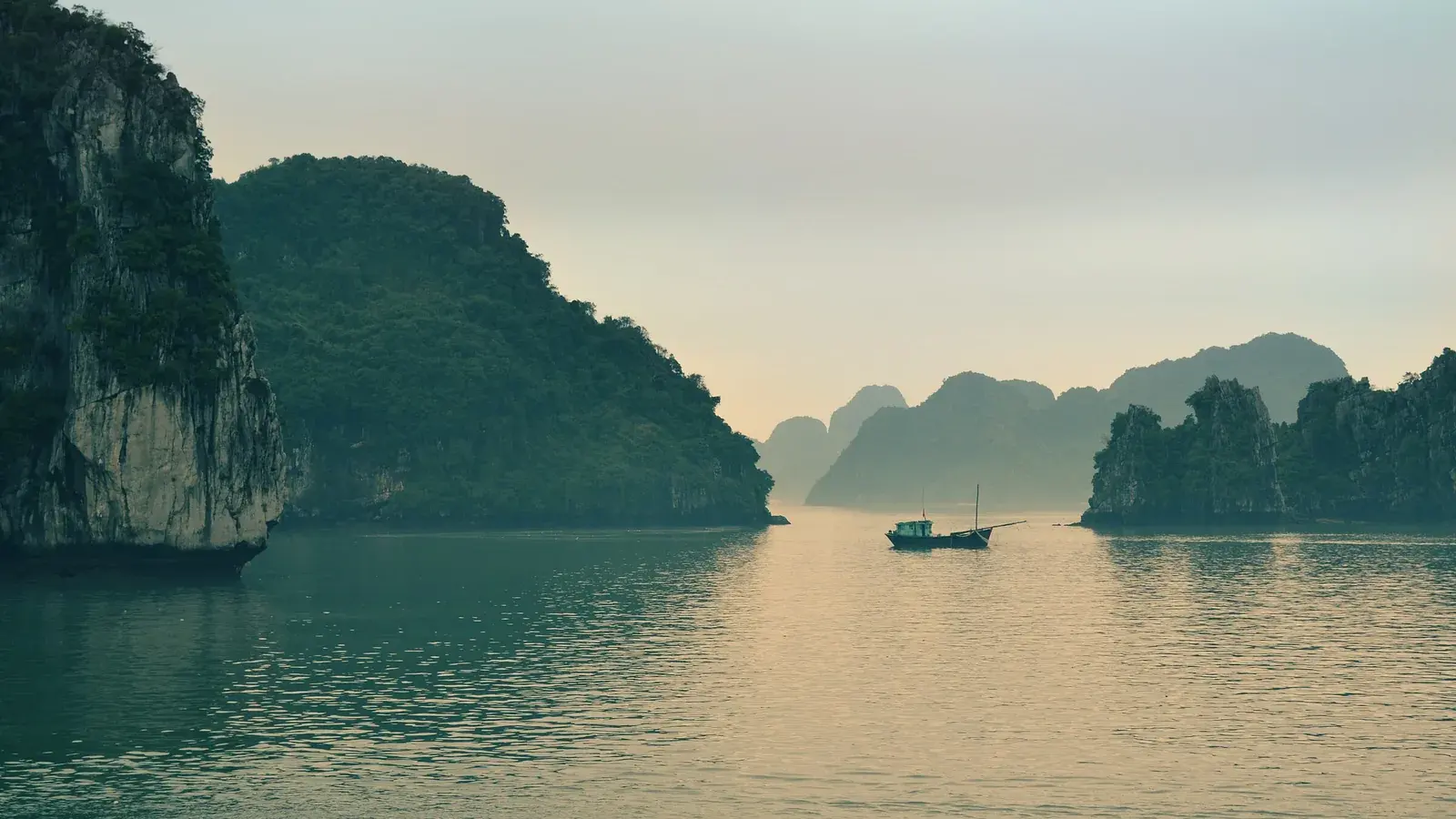 A wooden junk anchored between limestone karsts in a calm bay at dawn
