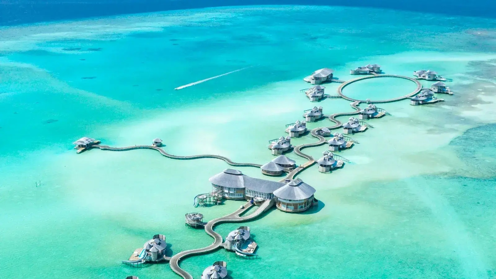 An aerial view of thatched overwater villas linked by curving boardwalks above a turquoise atoll