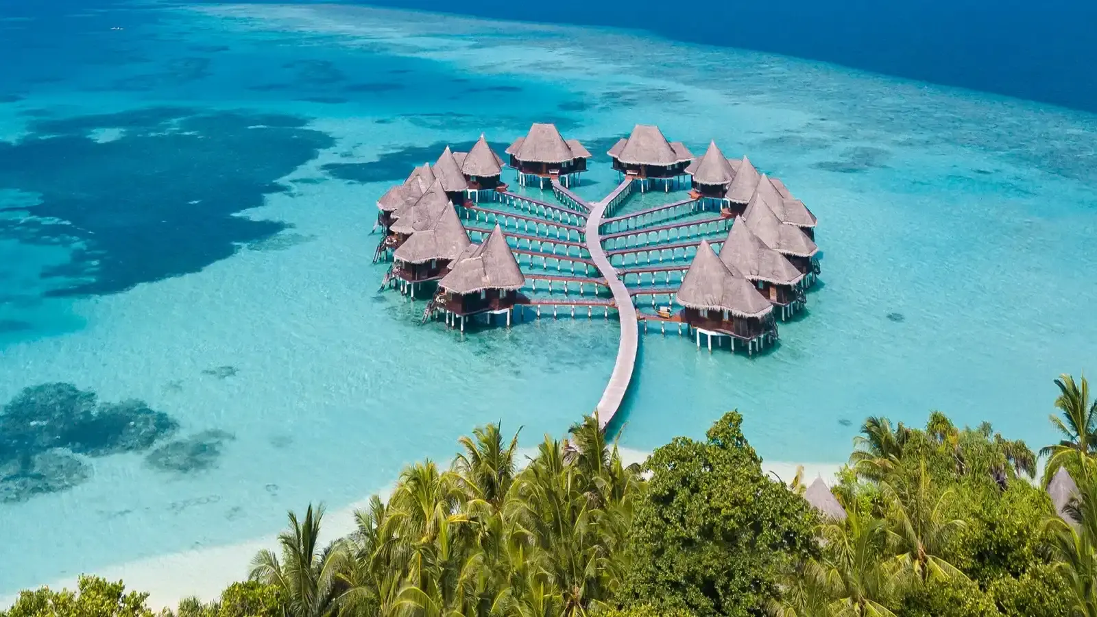 An aerial view of two rows of overwater bungalows reaching out across a pale lagoon