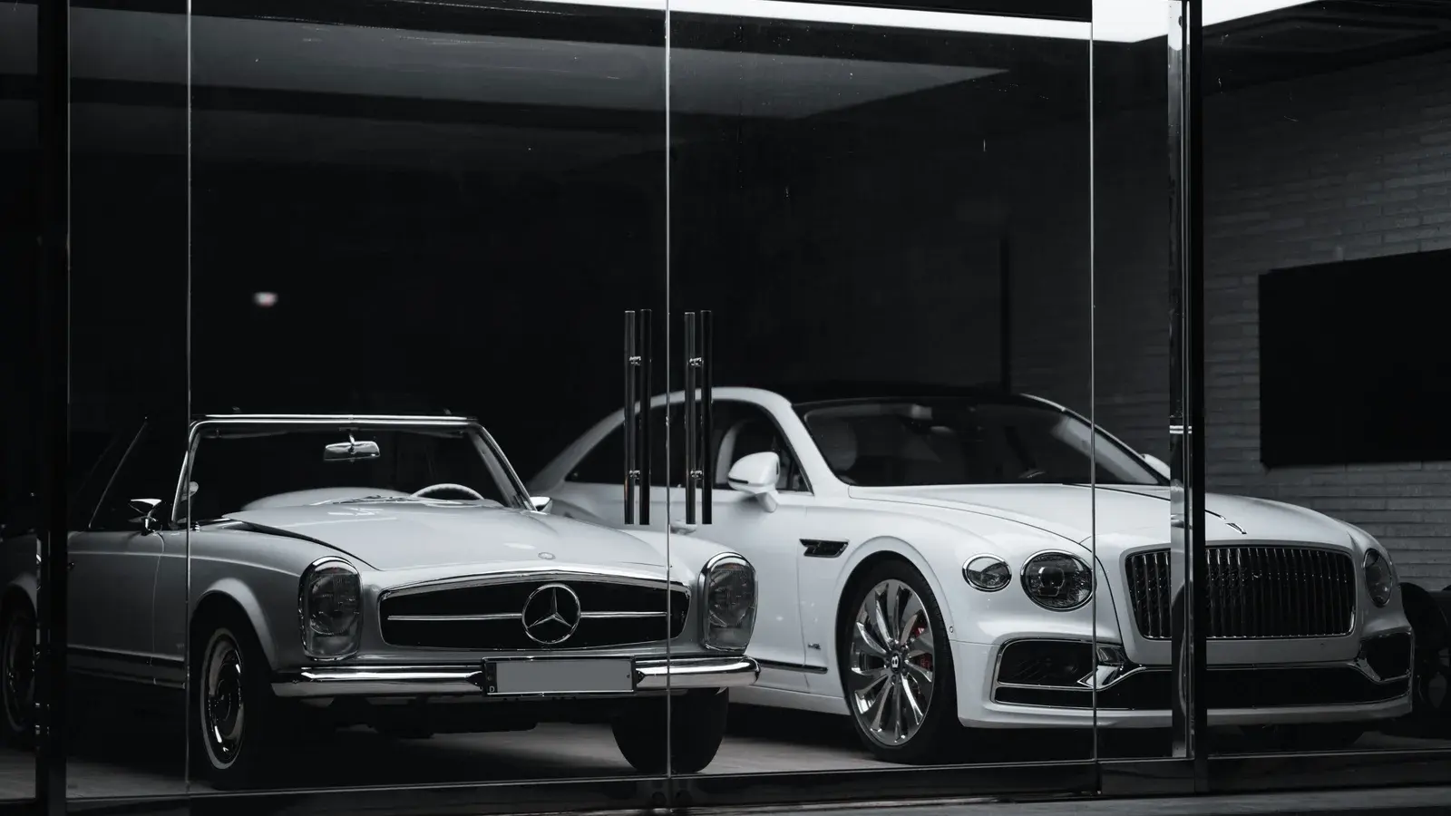 A classic silver roadster and a white convertible behind showroom glass