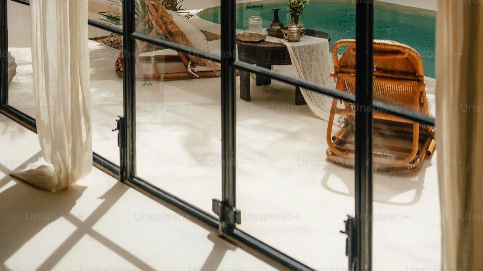 A rattan lounger and a pool terrace seen through steel-framed glass doors