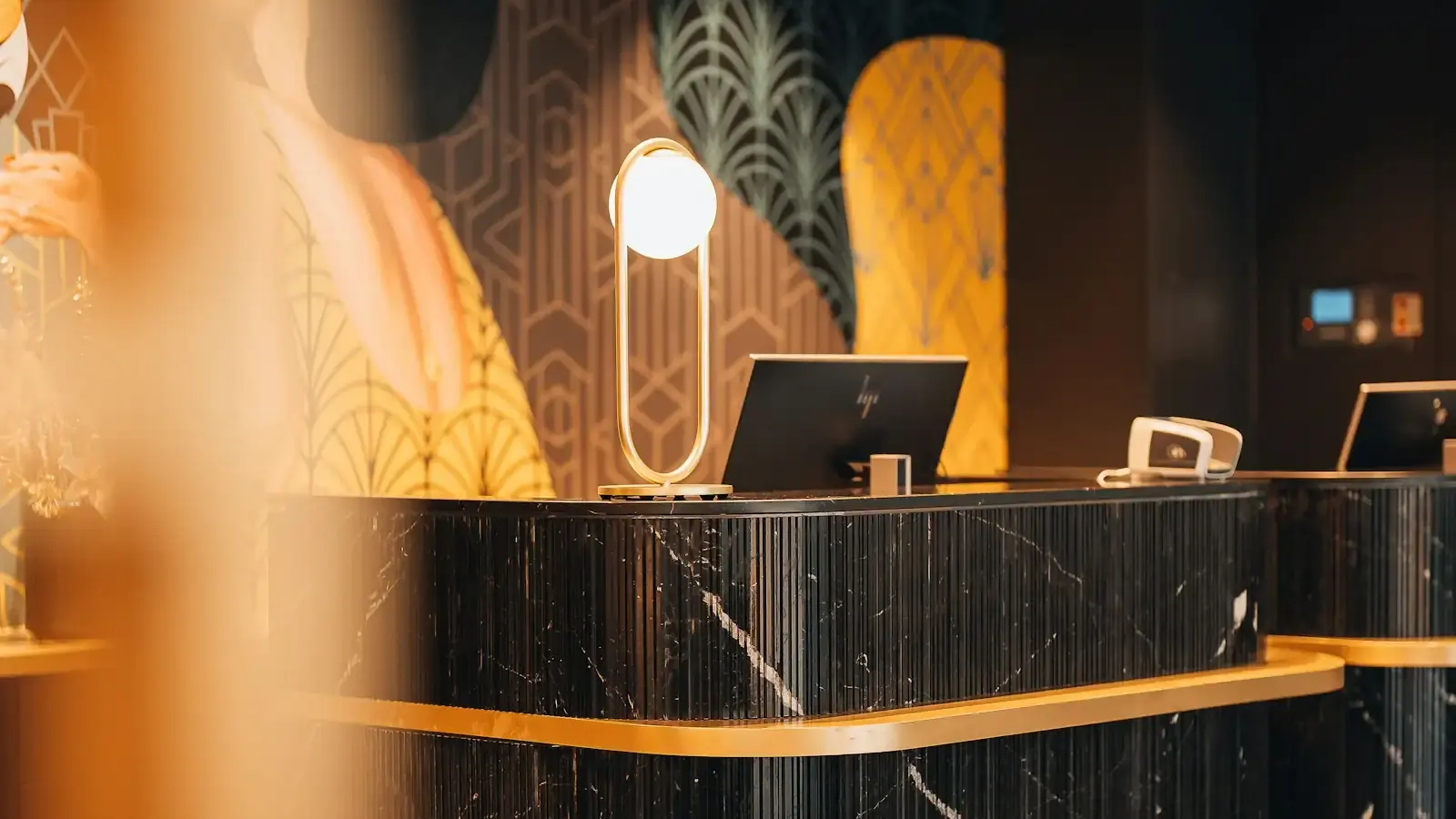 A black marble hotel reception desk with gold trim, a brass table lamp and a monitor, a patterned mural behind