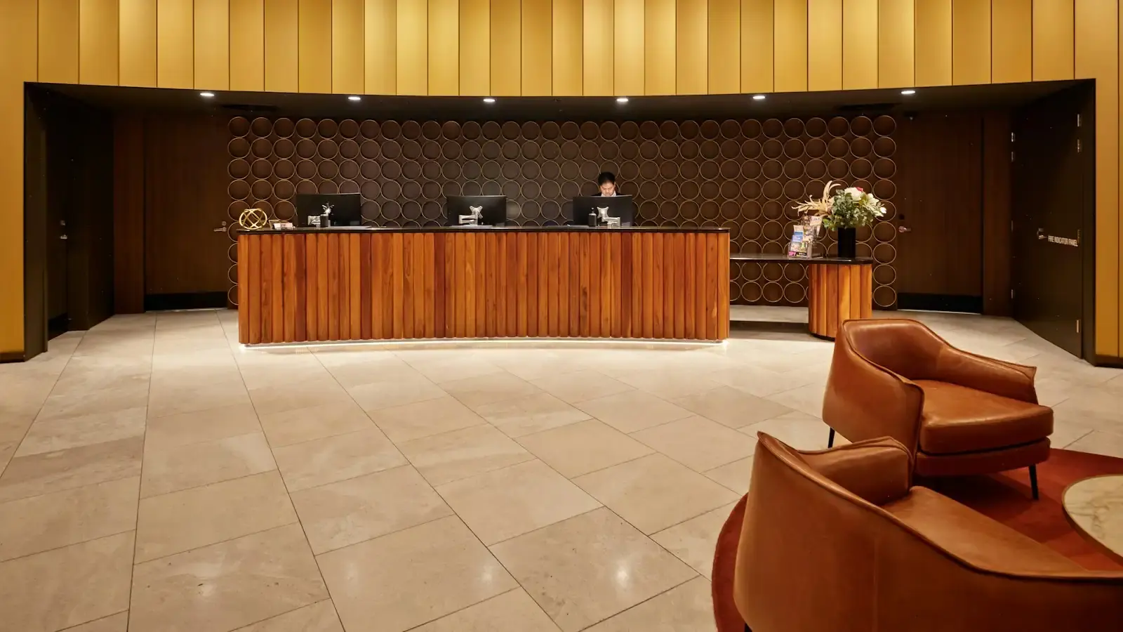A curved wooden hotel reception desk with three monitors in front of gold wall panels, leather armchairs in the foreground