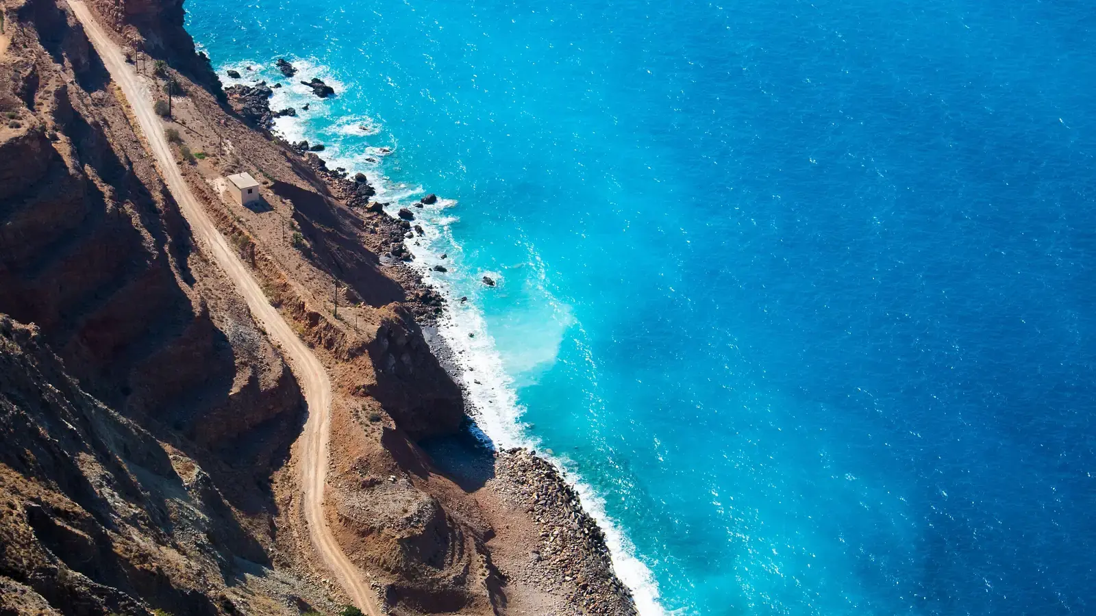 A dirt road cut along a red cliff high above a turquoise sea