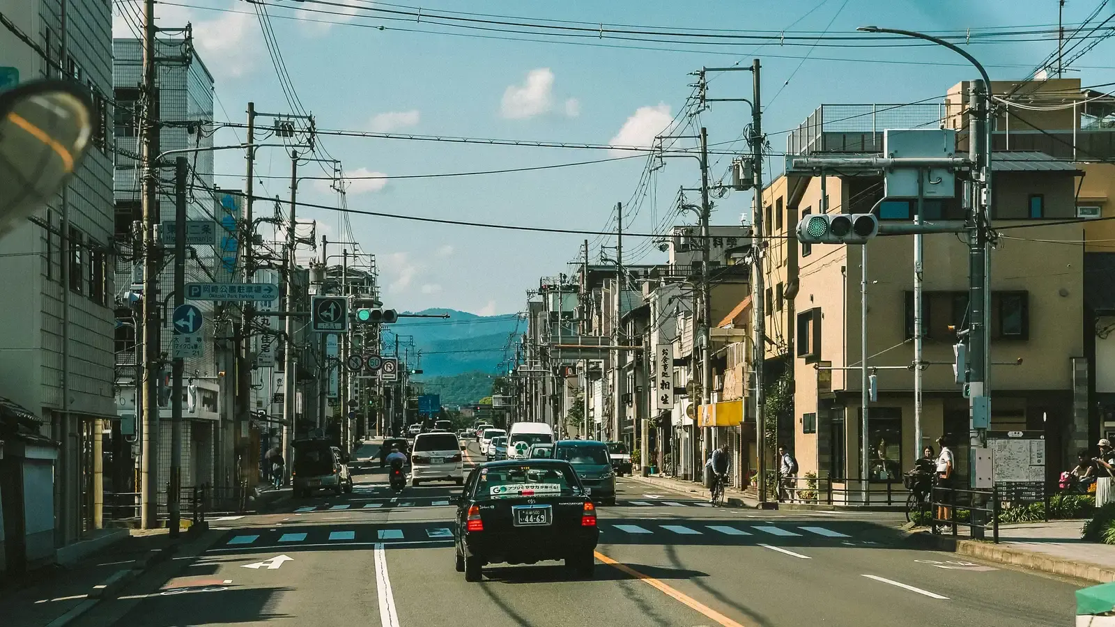 A car ahead on a Japanese street, power lines overhead and mountains beyond the rooftops