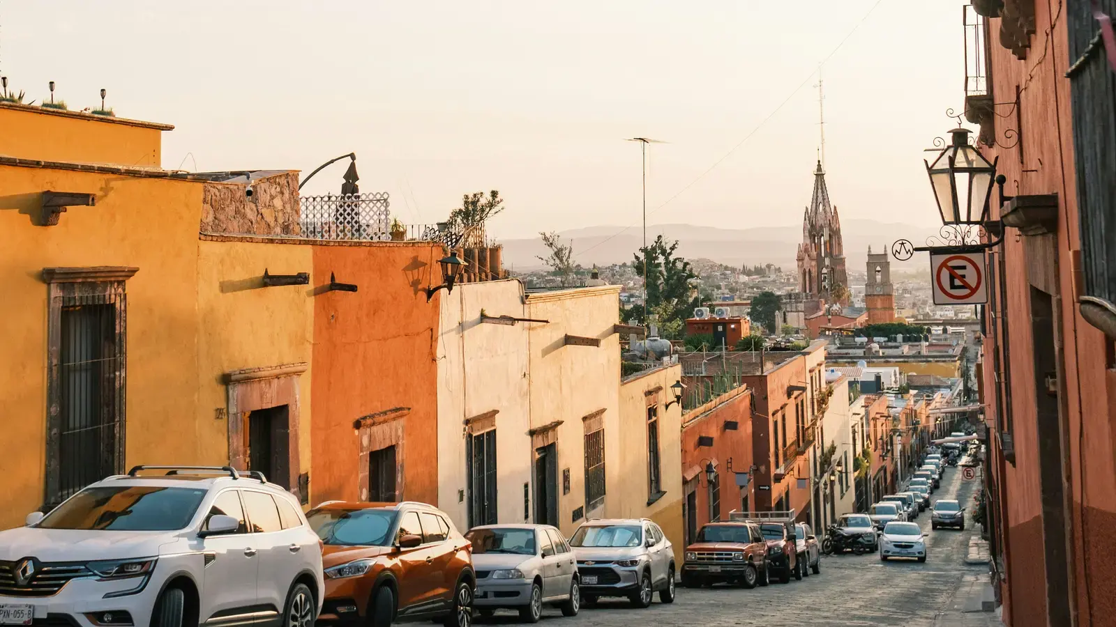 Cars parked along a colonial street in a Mexican hill town, a church spire on the skyline beyond