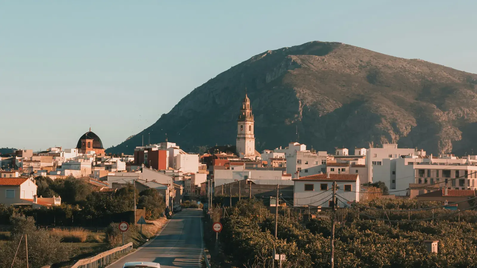 A white car on the road into a Spanish town, a church tower below a bare mountain