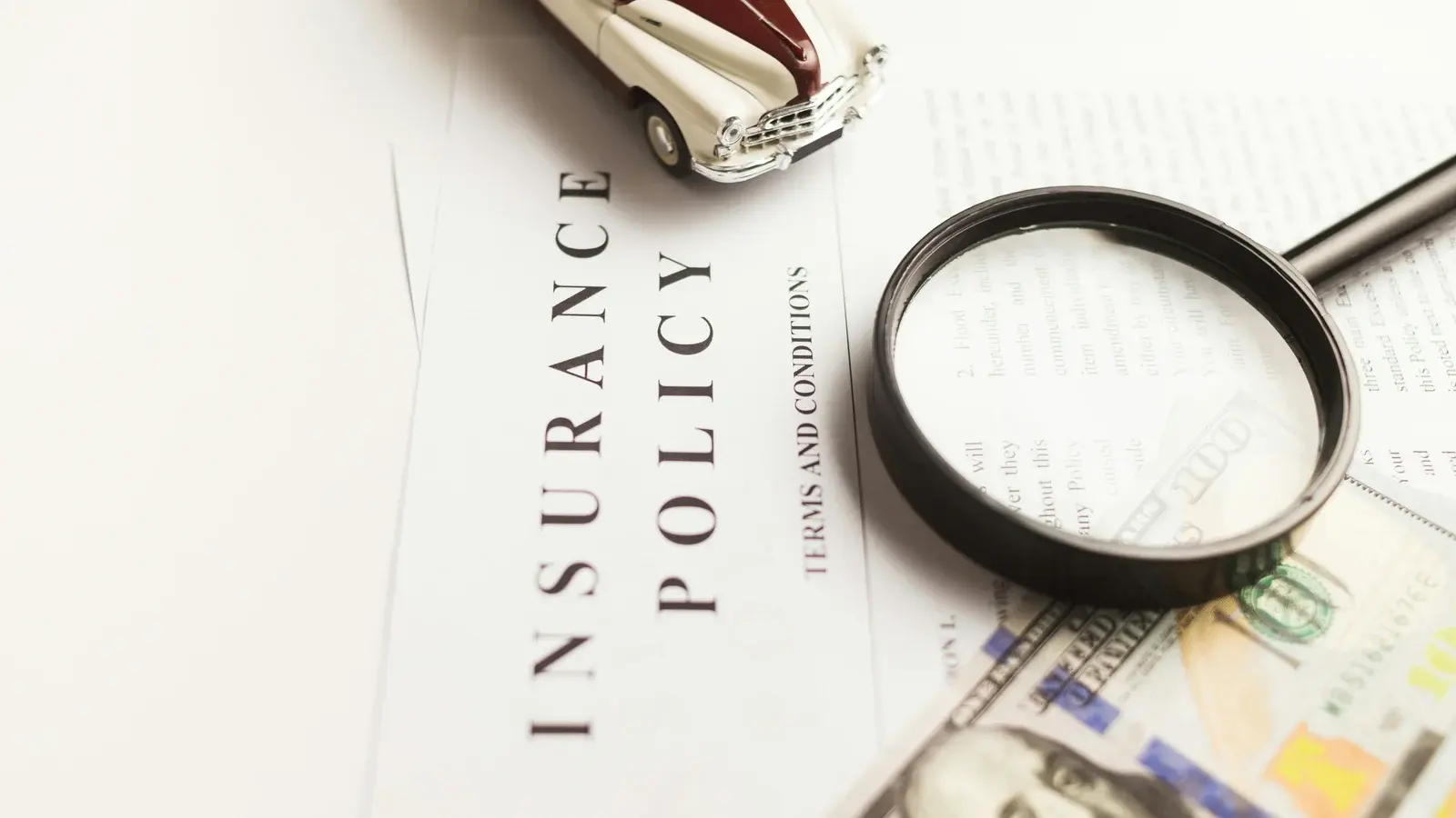 An insurance policy and its terms and conditions under a magnifying glass, a model car and banknotes beside them
