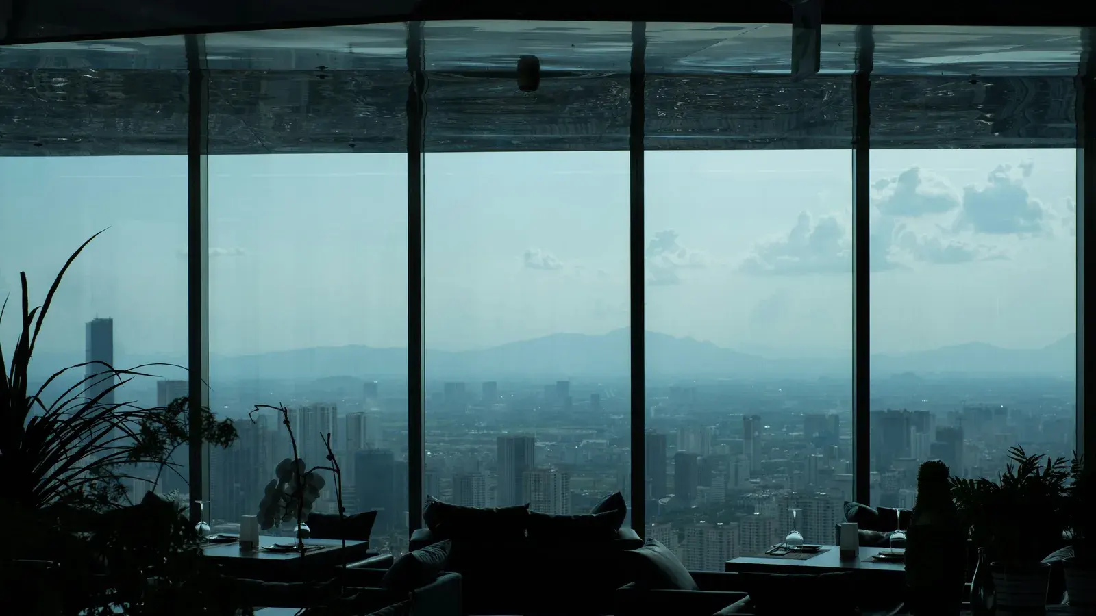 A dark restaurant terrace high above a hazy city ringed by mountains