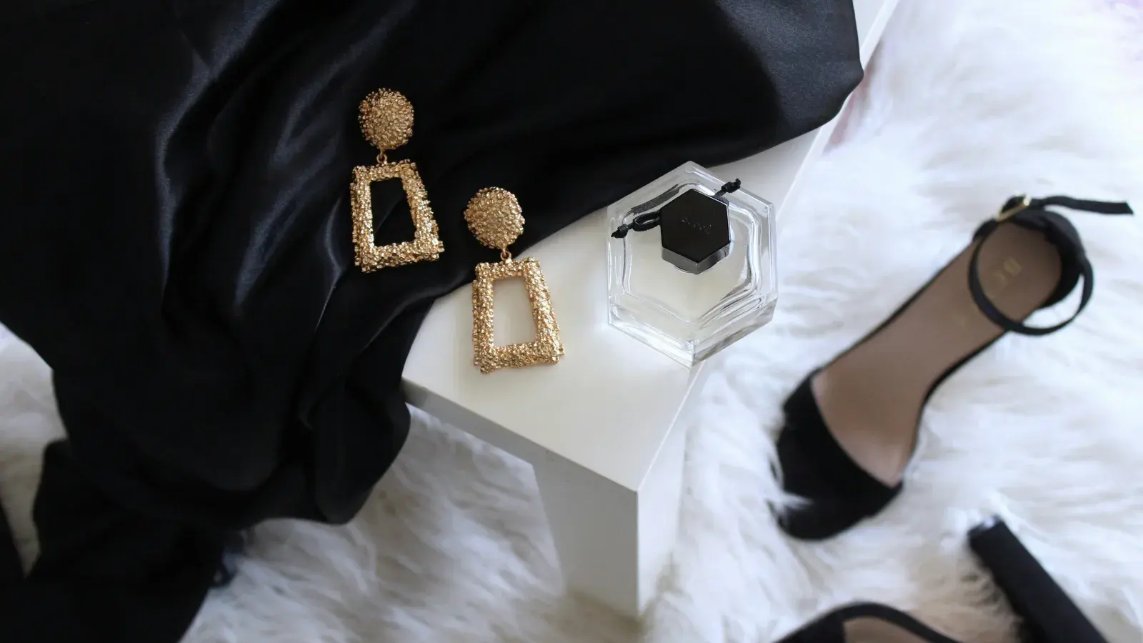 Gold earrings, a perfume bottle and black heels arranged on pale fur