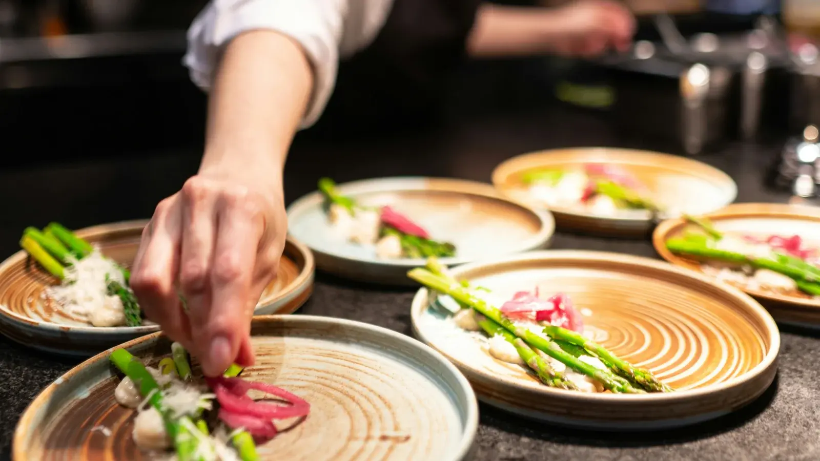A cook's hands finishing plates of asparagus and pickled onion on a kitchen pass