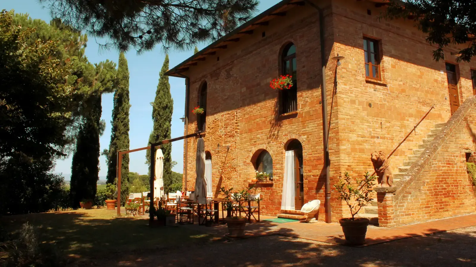 A brick Tuscan villa behind cypresses, its terrace laid with a table, chairs and folded parasols