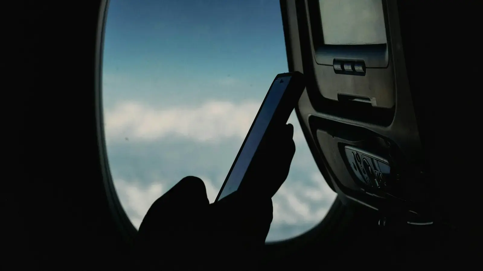 A silhouetted hand holding a smartphone in front of an aircraft window, clouds outside