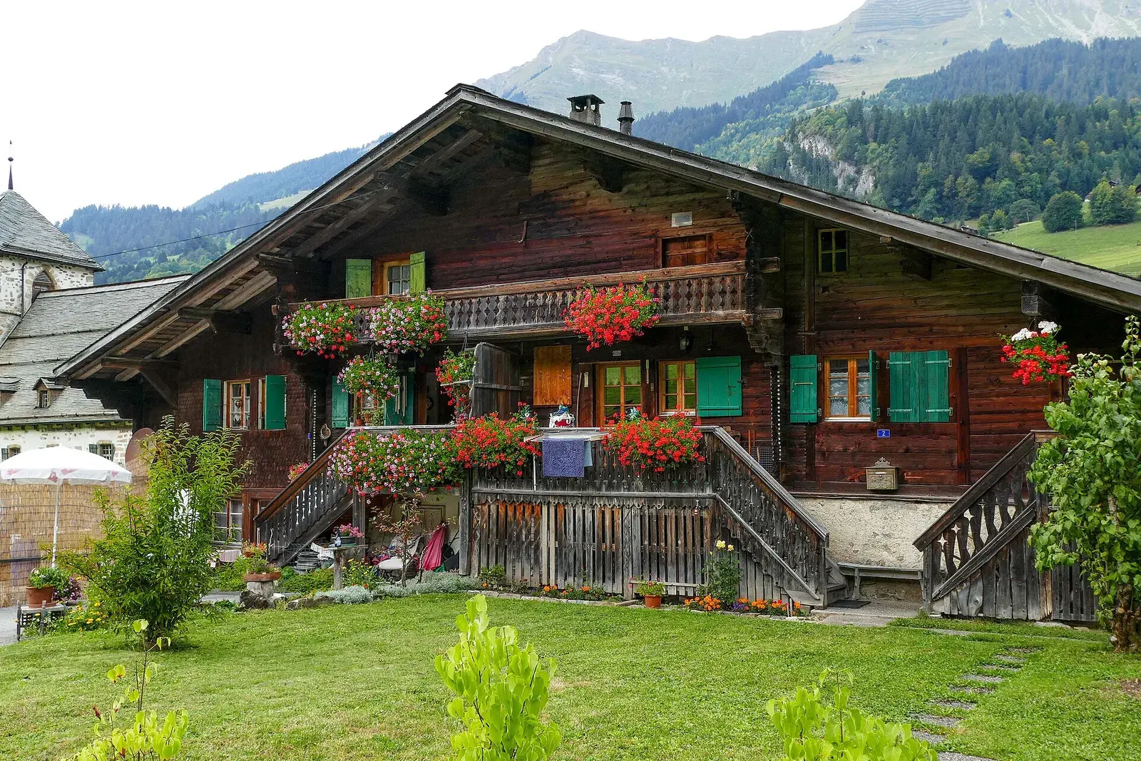 A timber alpine chalet hung with geraniums below a wooded mountainside
