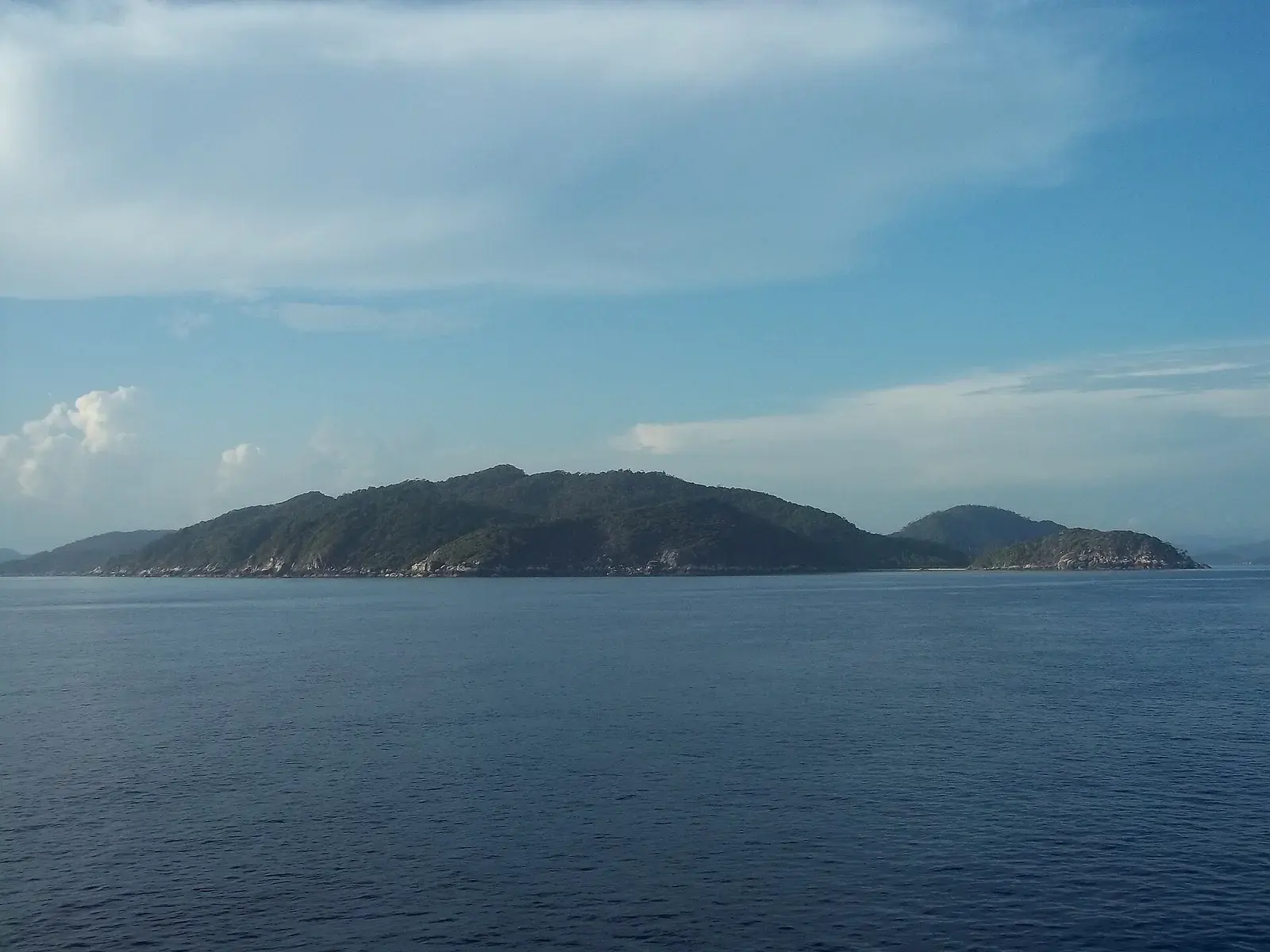 A long forested island seen at eye level from the water, its rocky shore under a partly clouded sky