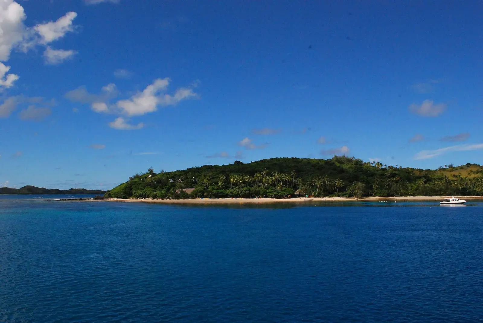 A forested island with a strip of pale beach, a small white boat lying off its far end