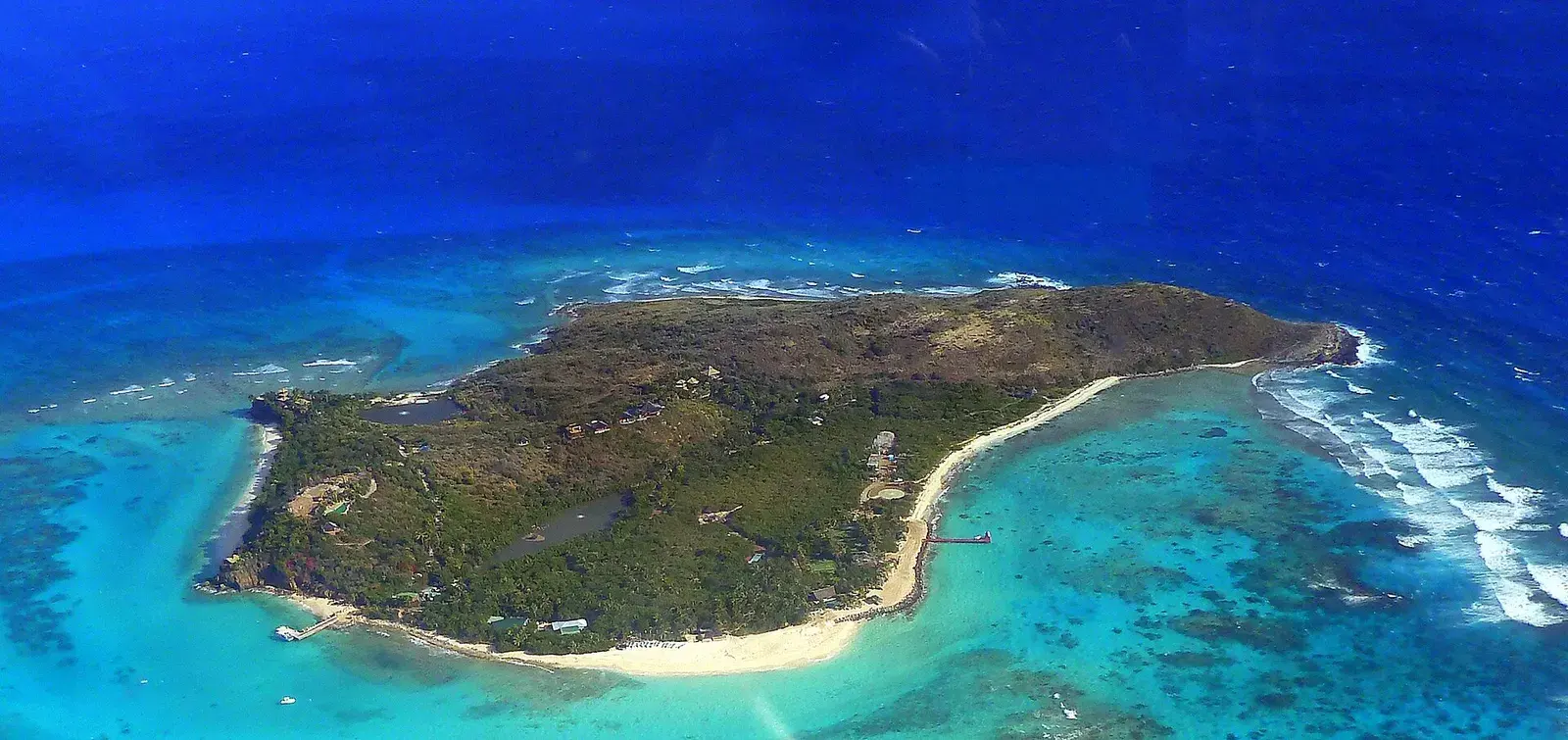 An aerial view of a low wooded island edged with white sand, jetties on both shores and reef breaking offshore