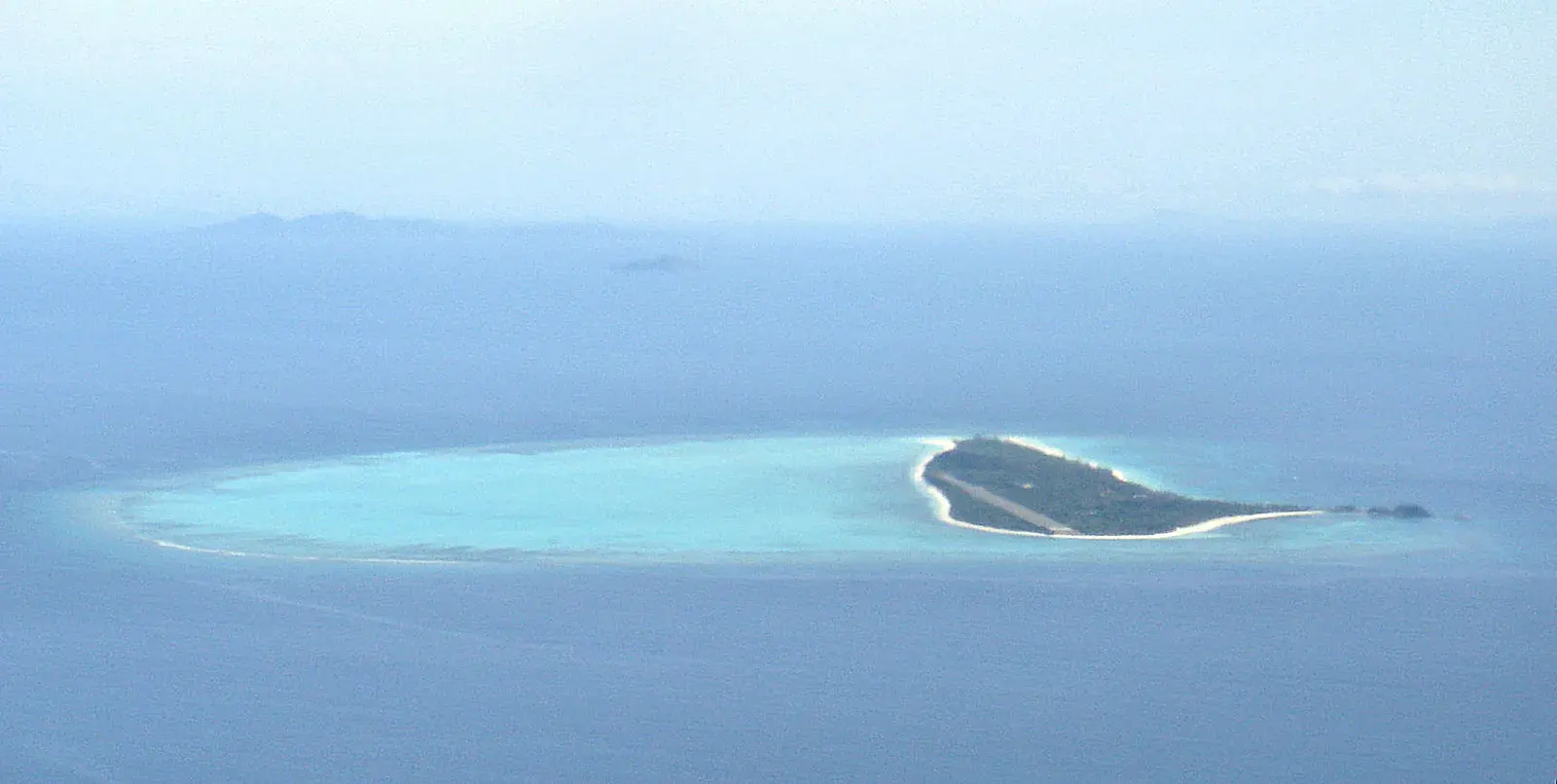 A distant hazy aerial of a low island and its pale lagoon in a wide expanse of blue sea