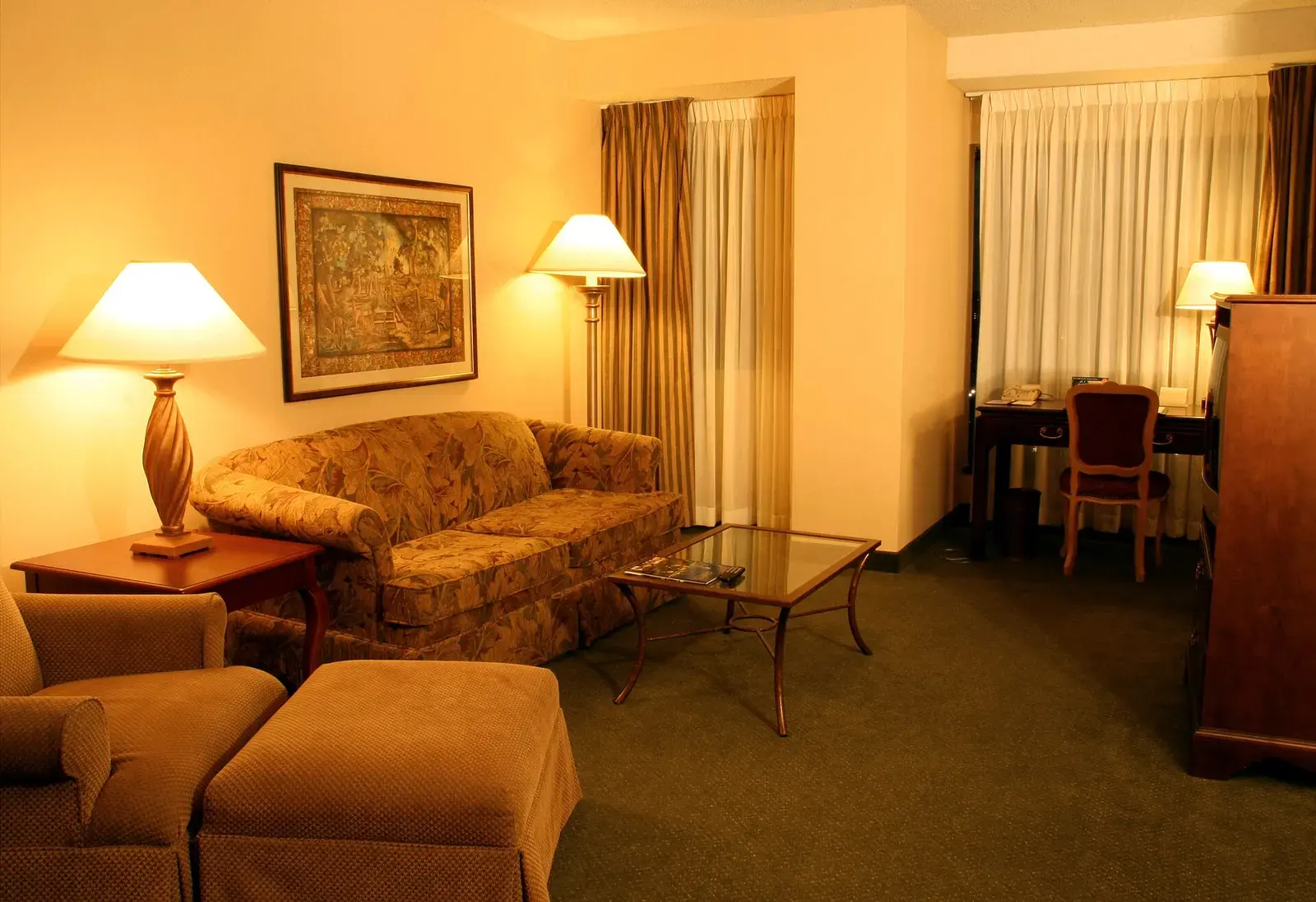 A hotel suite sitting room with lamps lit, a sofa facing a writing desk