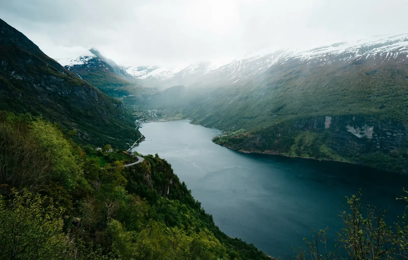 A fjord seen from high on a wooded mountainside, snow-streaked peaks and low cloud above a village at the head of the water