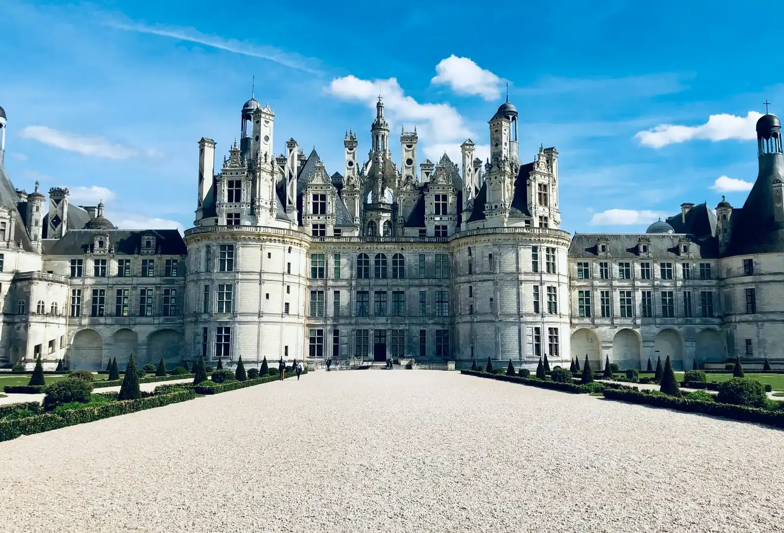 A French Renaissance chateau with round corner towers and a roofline crowded with chimneys and turrets, seen down a wide gravel walk between clipped hedges