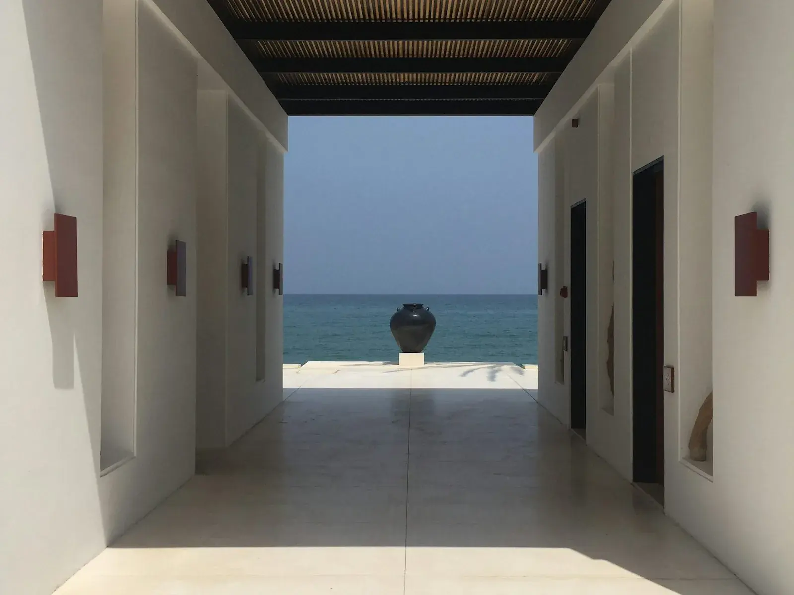 A white-walled resort passage opening onto turquoise sea, a dark ceramic urn on a plinth at the threshold