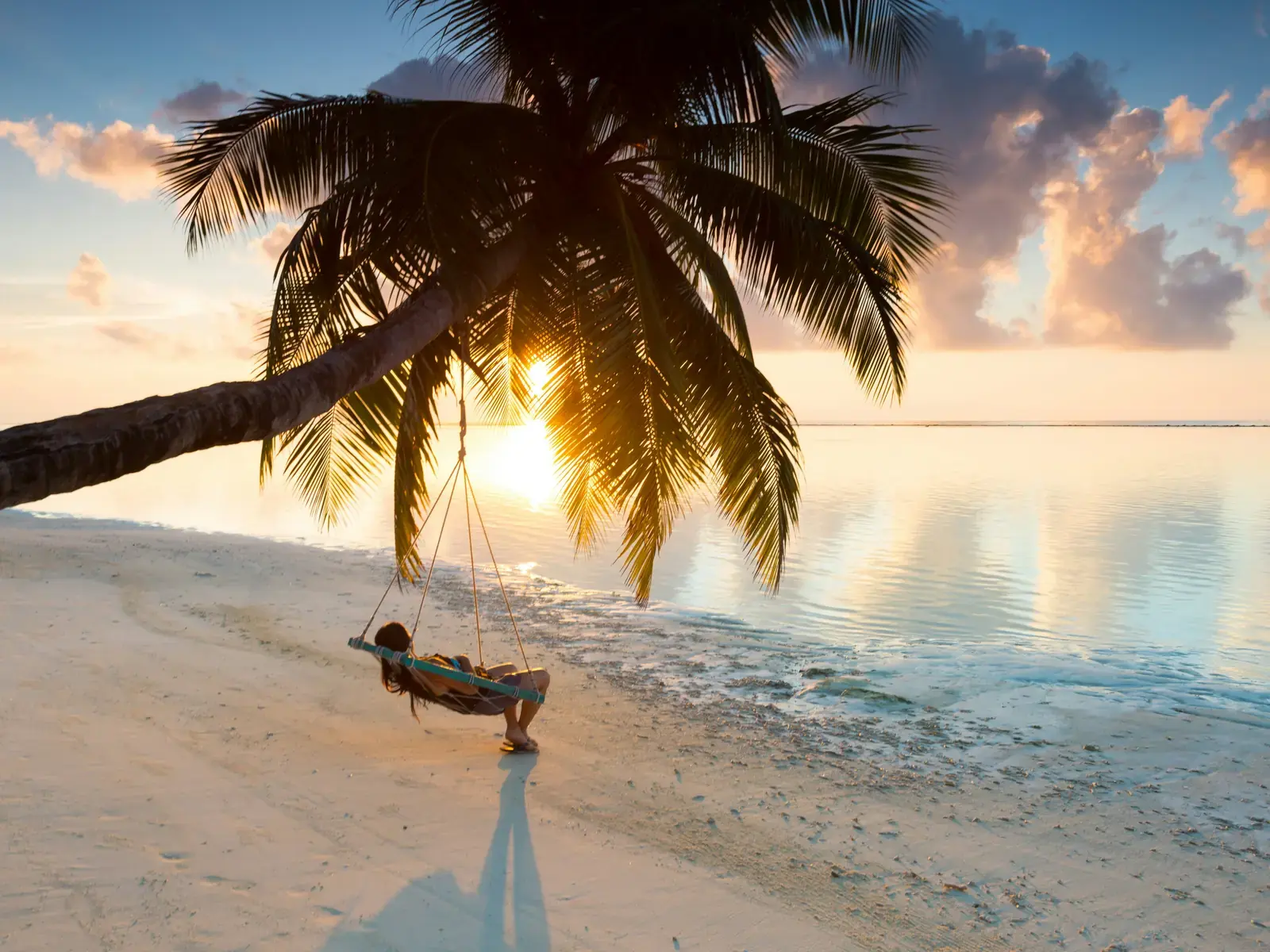 A woman lying on a rope swing hung from a leaning palm on white sand, the sun low over a flat lagoon