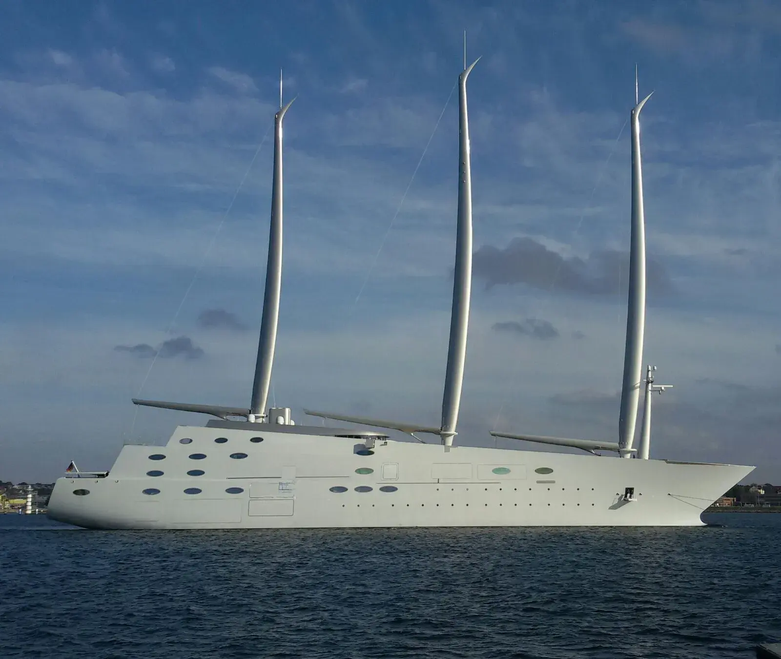 A three-masted white sailing yacht lying off a low coastline