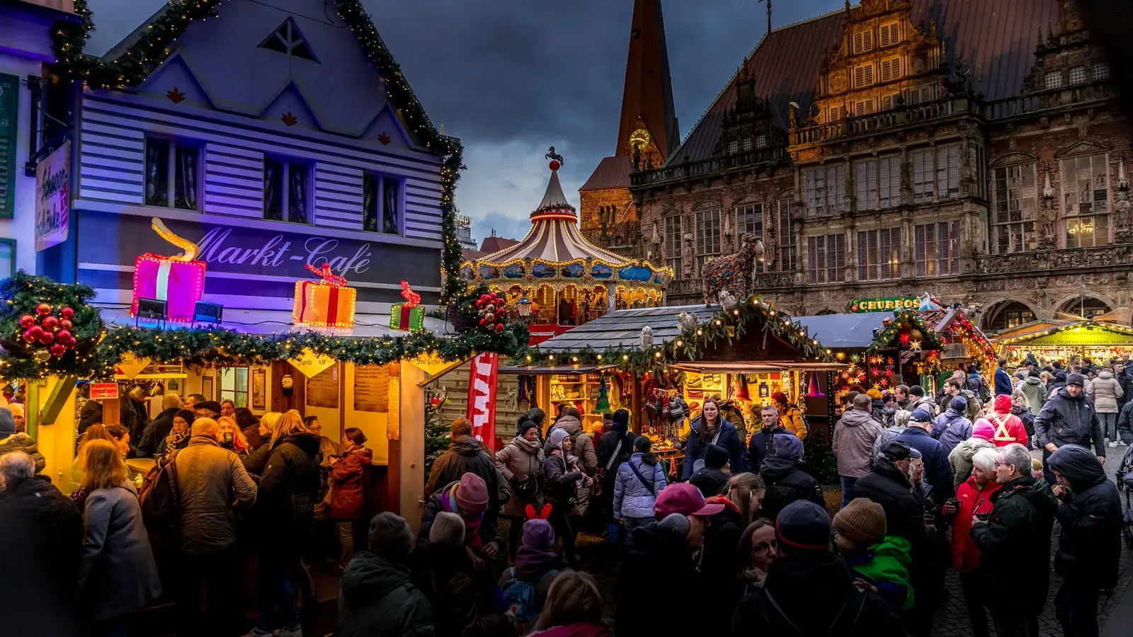 A crowded Christmas market at dusk, lit stalls and a carousel in front of a Gothic town hall and church spire