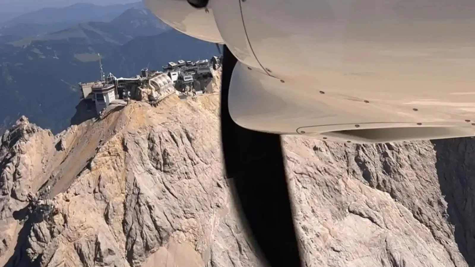 The cowling and propeller of a small aircraft passing close to a rocky Alpine summit topped with buildings