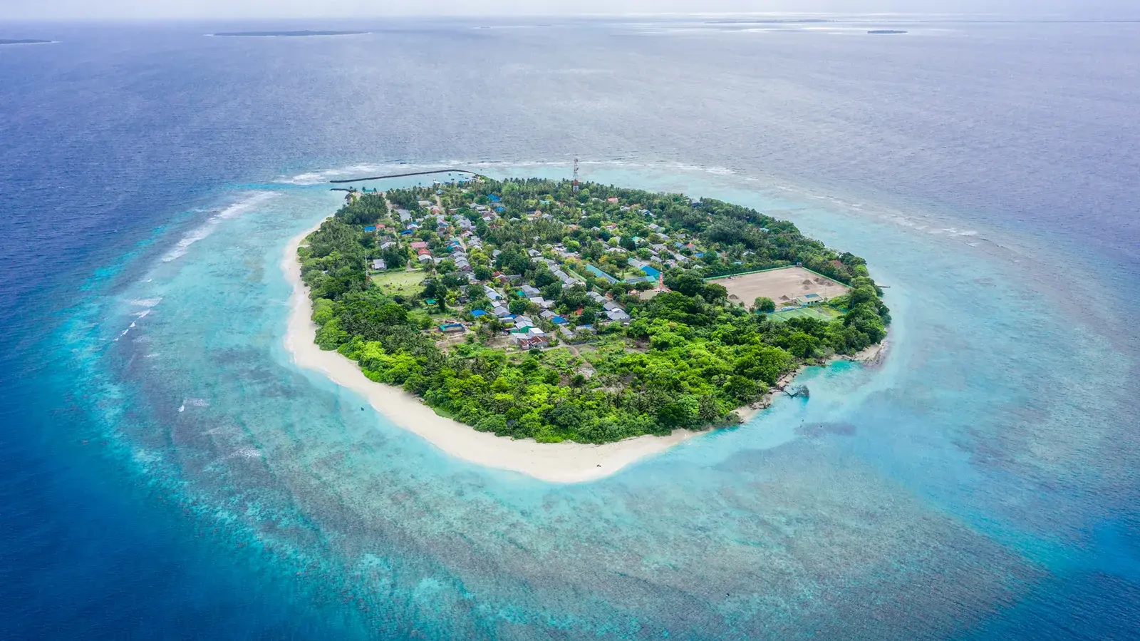An aerial view of a small inhabited island of low roofs and trees, ringed by white sand and shallow reef
